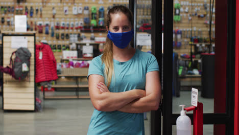 comerciante caucásica con máscara facial en la puerta de una tienda de deportes con los brazos cruzados