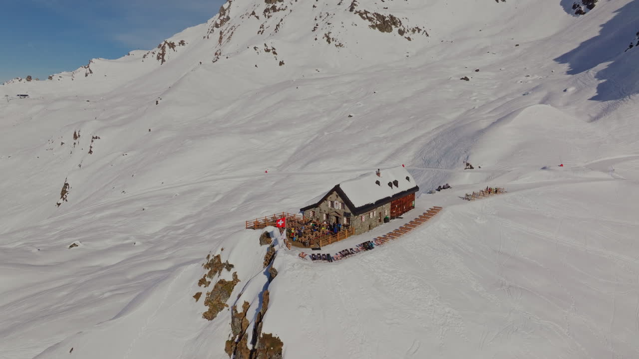Aerial orbit of Cabane Mont Fort, mountain hut in Verbier, Switzerland, beneath the iconic Bec des Rosses on a clear alpine day.