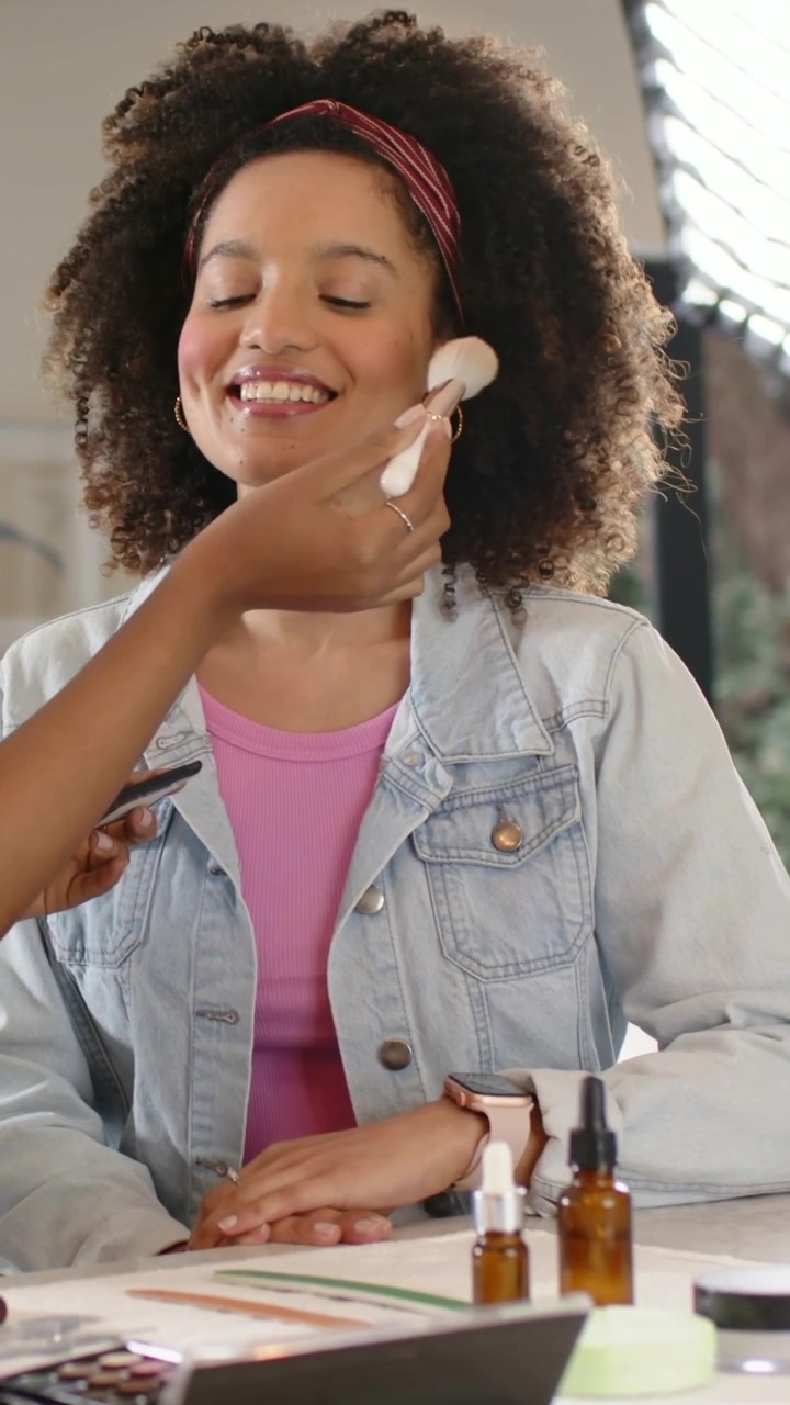 Vertical video: Smiling woman getting makeup in studio, pink top, artist bringing brush, palette