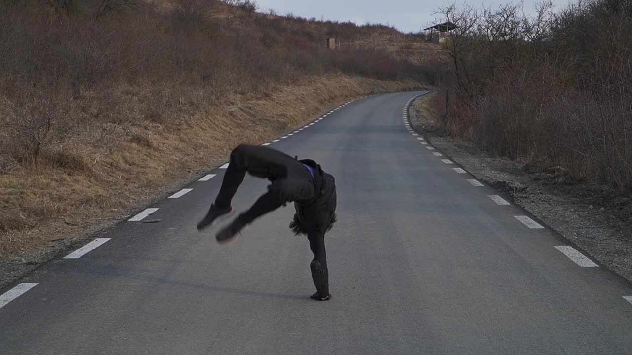 Acrobatic man in black performs a one-hand spin followed by a shooting gesture on an empty road. Perfect for action scenes, dance, or dynamic storytelling clips