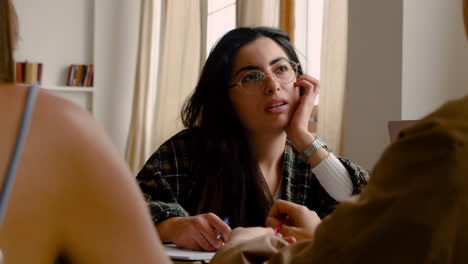 camera focuses on a brunette girl with eyeglasses of a study group writting sitting at table and talking with othes mates