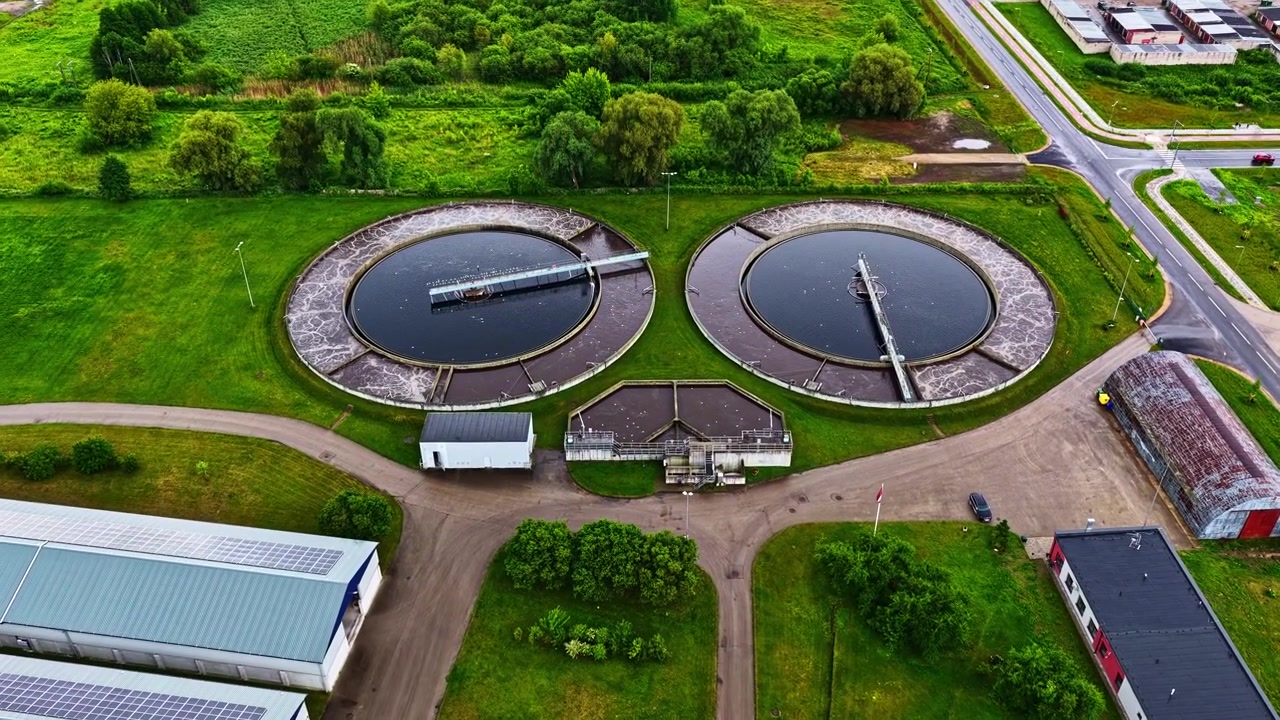 Two Large Circular Clarifier Basins Operate within a Treatment Plant Surrounded by Roads, Buildings, Ponds, and Green Landscapes - Drone Flying Forward
