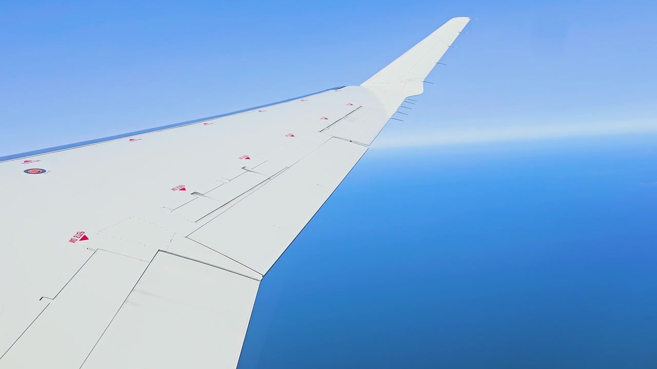 飛行機の窓から見た翼の景色、広大な外洋上空を遠くにかすみがかった澄み切った青空の中飛行する様子、民間航空の旅の情景、穏やかな空の旅のPOV映像、そして広がる海の水平線。