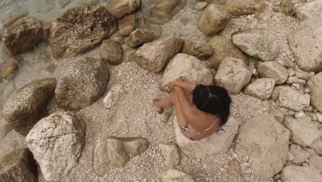 View-from-above-of-a-girl-sitting-on-a-white-rocky-beach-and-looking-at-the-sea