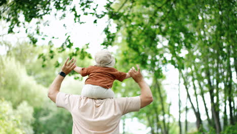 caucasian positive man holds a baby in a cap on his shoulders. father holds his son by the hands and runs in the park.