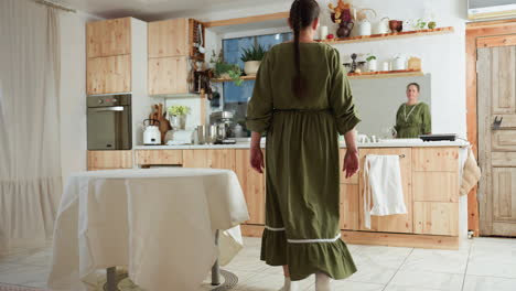 lady in long green dress with braid hairstyle walks into cozy wooden kitchen, decorated with indoor plants, hanging utensils, and white tablecloth-covered round table, ready to begin daily routine