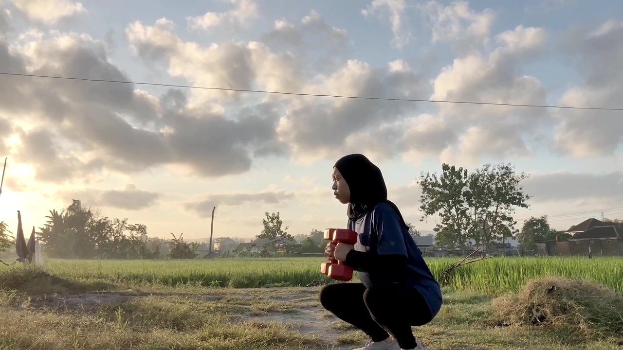 Video gerak lambat profil samping seorang wanita Muslim Asia berhijab yang melakukan squat dengan dumbel di luar ruangan. Dia berolahraga di lapangan saat langit matahari terbit yang indah.