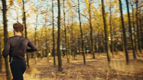 a male athlete with curly hair and a beard in a black sports uniform runs quickly through an autumn forest with fallen leaves and dry grass