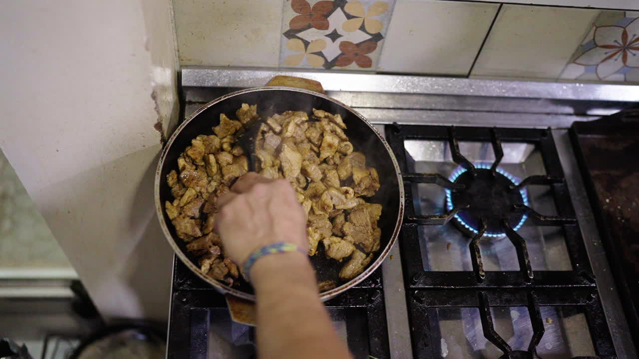Local pork cuisine preparation closeup clip on large black pan inside household kitchen, with someone's hand stirring it over.