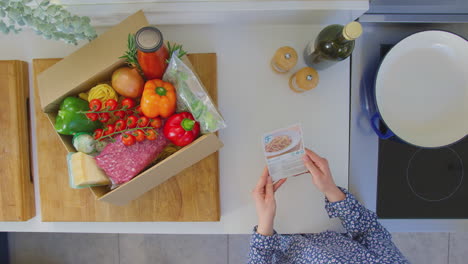 Vista-Desde-Arriba-De-Una-Mujer-Con-Tarjetas-De-Recetas-Y-Una-Caja-De-Ingredientes-Entregados-Planificando-La-Comida---Filmada-En-Cámara-Lenta