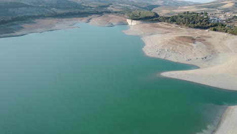 Aerial-shot-over-the-turquoise-water-and-sand-of-a-reservoir-with-a-dam-in-the-background