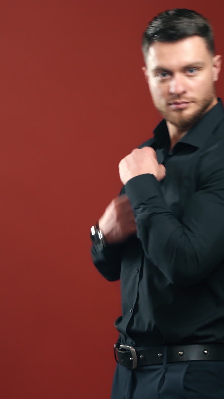 Confident businessman in stylish wear. Handsome man with beard posing on camera in dark red studio. Portrait of young brunette male in black shirt. Vertical video