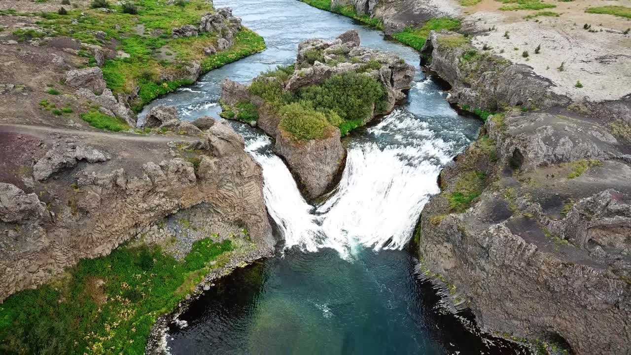 Hjálparfoss waterfall is creating white, foamy water as it plunges down moss-covered rocky cliffs into a turquoise pool in Iceland, showcase raw beauty of Icelandic nature, drone pulling out shot