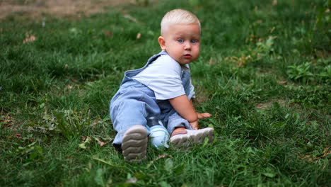 adorable child in a jeans romper sitting in the green grass in summer. little toddler boy playing outdoors.