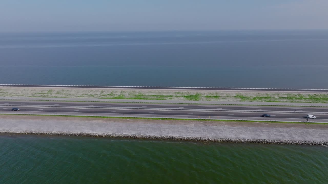 Drone zoom in flying over the Afsluitdijk highway dividing the Wadden Sea and IJsselmeer under clear blue sky