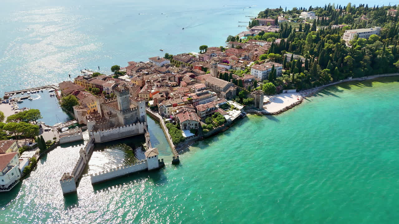 Vista aérea de drone do Castelo Scaligero em Sirmione, cercado pelas águas turquesa do Lago de Garda. Muros e torres medievais erguem-se acima do porto e da histórica cidade à beira do lago.