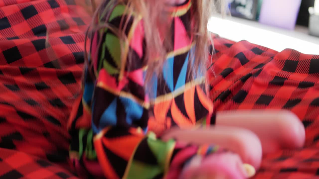 Little kid caucasian white child with long hair jumping on bed with red and black plaid blanket and falling on  knees wearing a colourful neon colored sweater - in Cinema 4k (30fps slowed from 60fps).