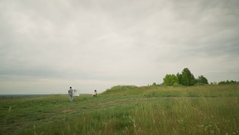 a back view of an artist wearing a hat and checkered shirt, deeply focused on painting on an easel in a lush grass field under a cloudy sky. a woman in a hat and white gown sits calmly in a chair