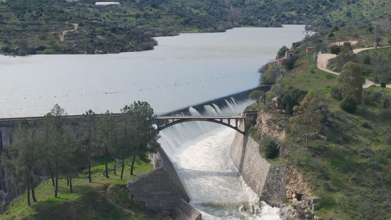 Descending drone flight in front of a dam overflowing through its spillway with impressive force. The falling water flows beneath a bridge located right over the roaring spillway.