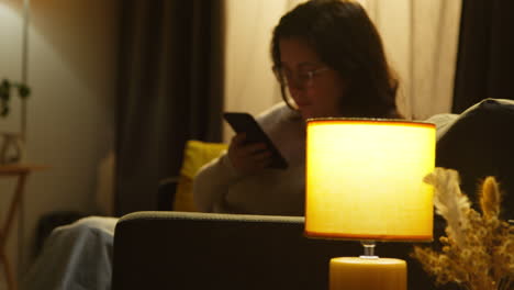 young woman at home sitting on sofa with mobile phone scrolling through internet or social media in the evening 4