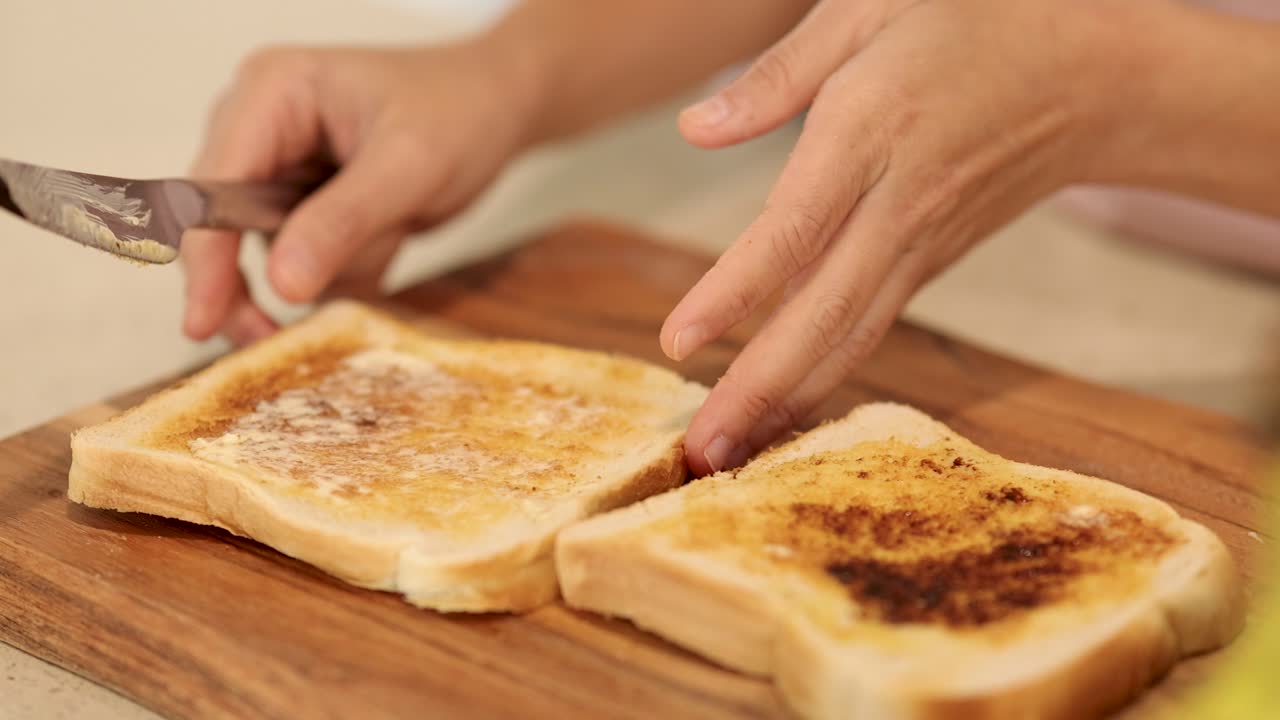 Hands spread Vegemite on bread slices using a knife on a wooden board in a well-lit kitchen