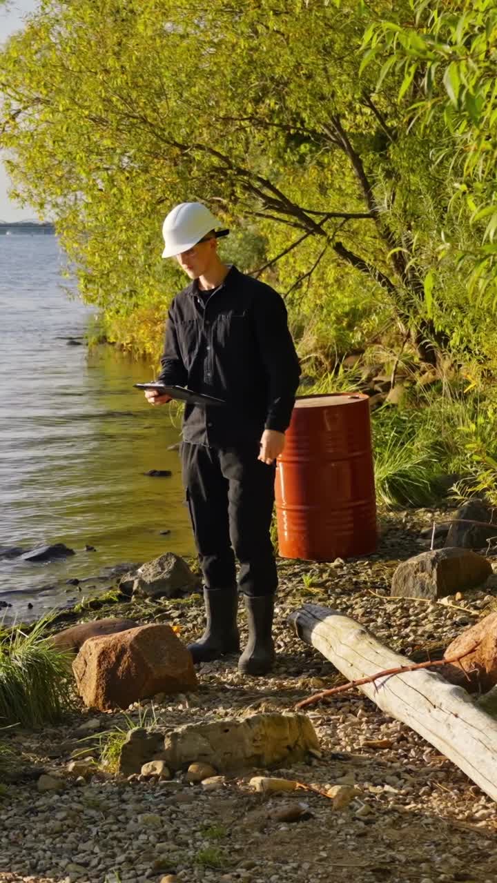 Inspector in black clothes and rubber boots writes notes near polluted lakeside area, vertical