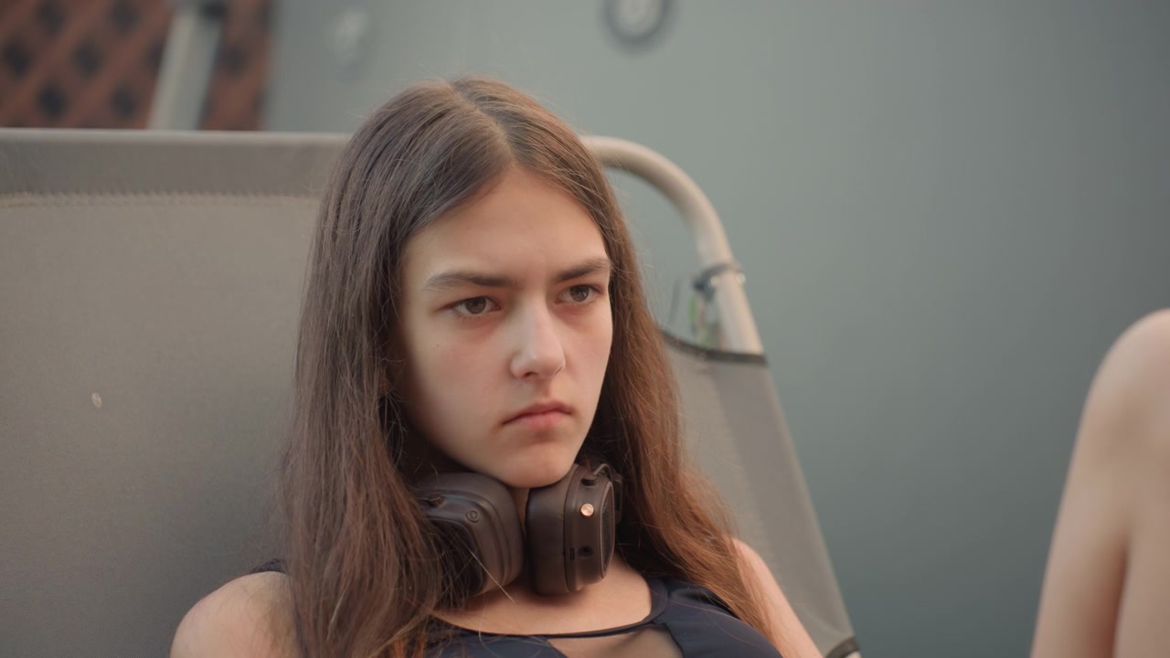Una giovane donna si rilassa su una sedia a sdraio grigia, tenendo e usando il suo smartphone mentre indossa grandi cuffie over-ear. La donna è vestita con un top scuro con dettagli intagliati, circondata da una luce soffusa.