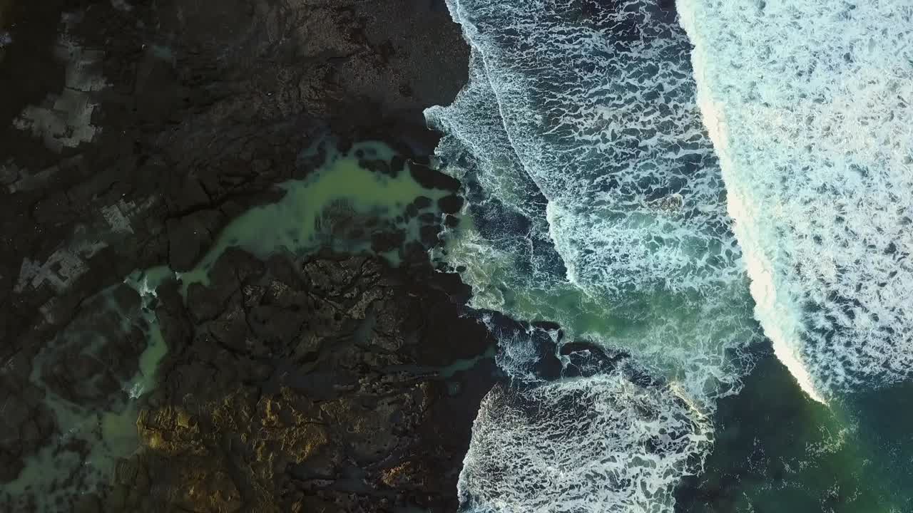 Tanah lot coastline, ocean waves crashing against dark rocky shore, aerial view