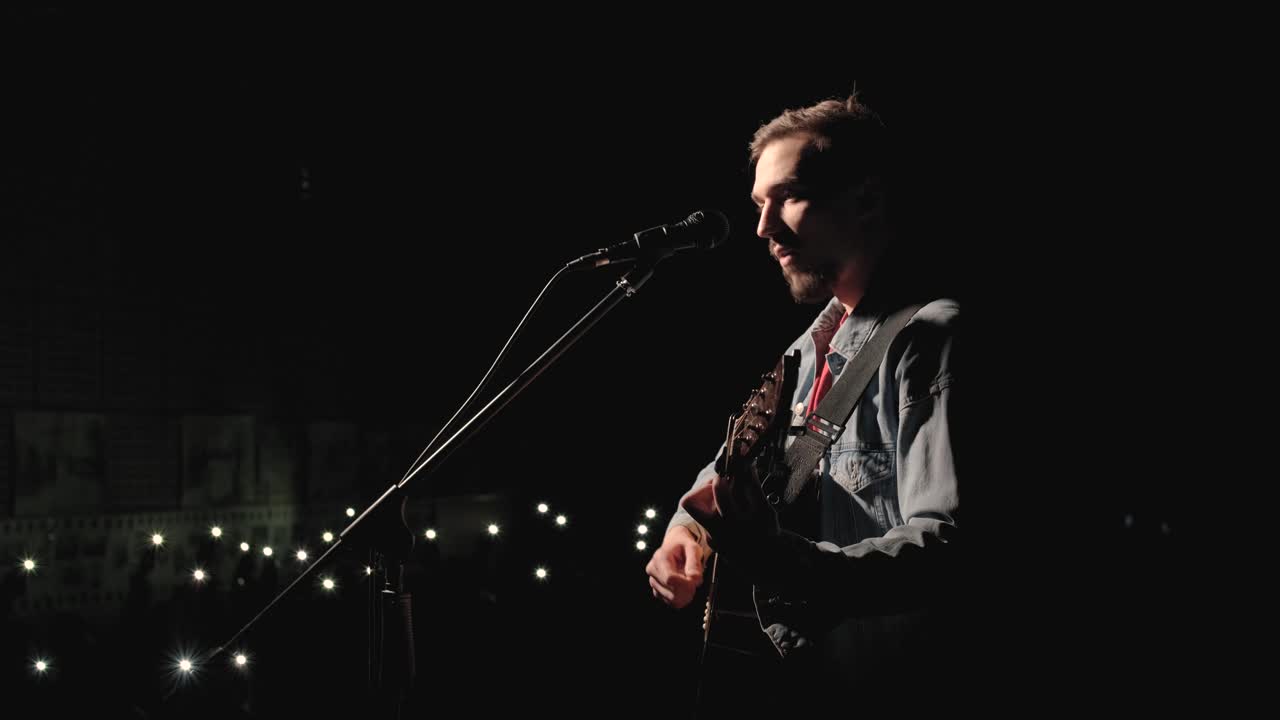 한 뮤지션이 큰 무대에서 기타를 연주하고 있으며, 관객들은 배경에 불을 들고 있습니다. 관객실은 아름다운 박이는 불빛으로 가득 차 있습니다.