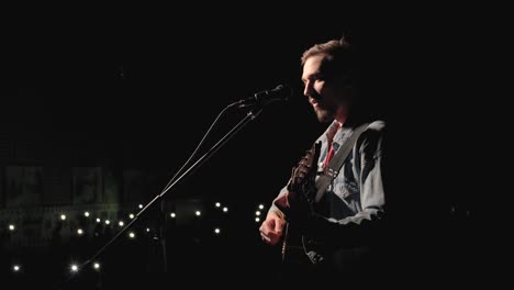 a musician is playing guitar on a large stage with audience members holding flashlights in the background. the auditorium is filled with beautiful flashing lights.