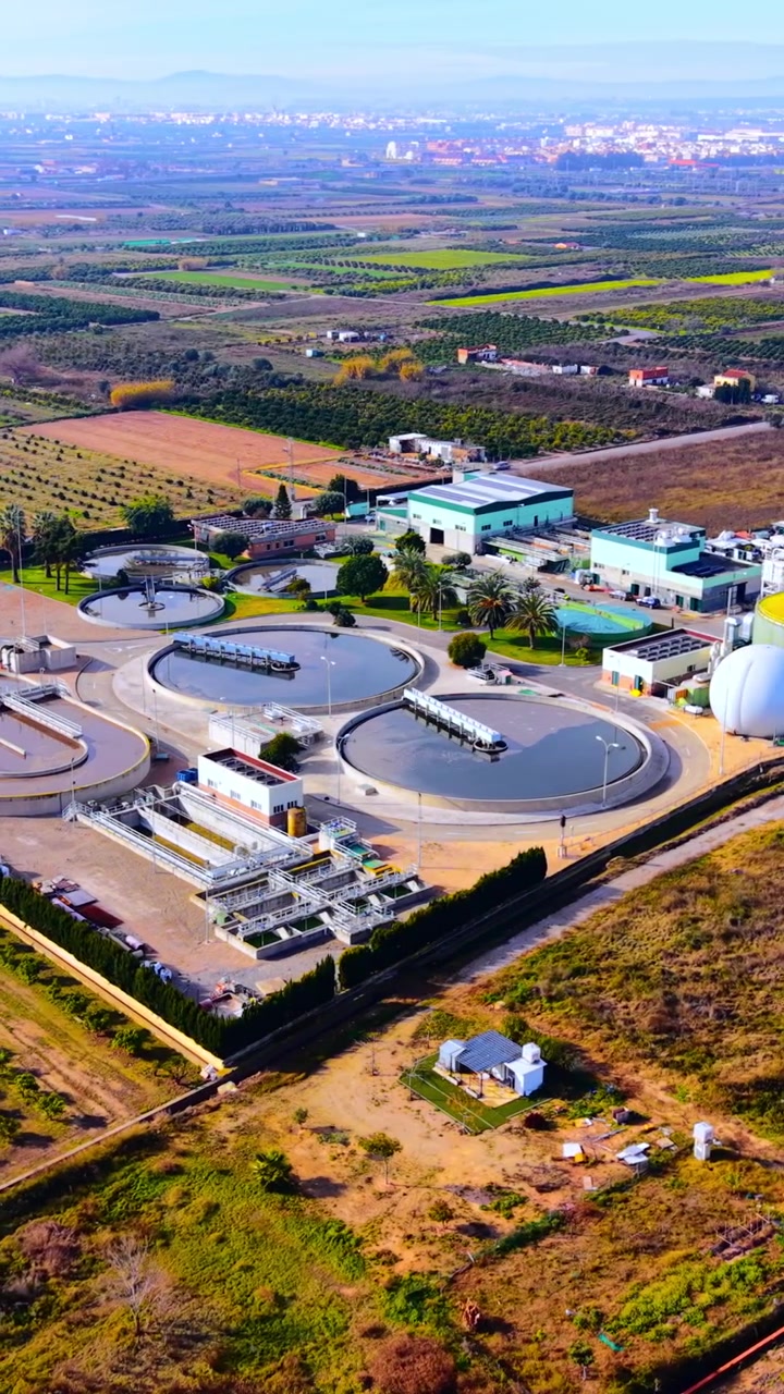 Planta de tratamiento de aguas residuales con tanques circulares. Clarificadores redondos, gasómetro de biogás, paneles solares en el techo, palmeras y huertos circundantes en una llanura agrícola. Vídeo vertical