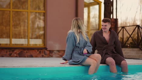 man and woman are resting in thermal bath in recreation complex sitting on edge of outdoor swimming pool