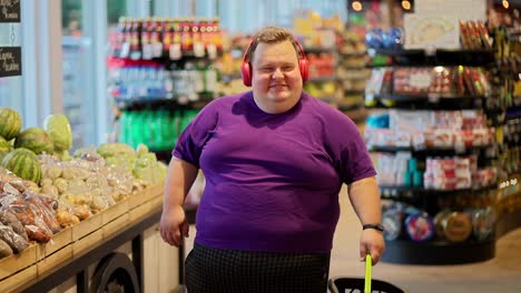 A-happy-man-in-a-purple-T-shirt-and-red-headphones-walks-through-the-supermarket-and-listens-to-music-while-shaking-his-head.-A-fun-trip-to-the-supermarket-while-listening-to-music