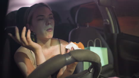 woman in black top eating doughnut while on the phone in parked car at night, appearing frustrated by the conversation, with a shopping bag visible in the backseat, under soft interior lighting