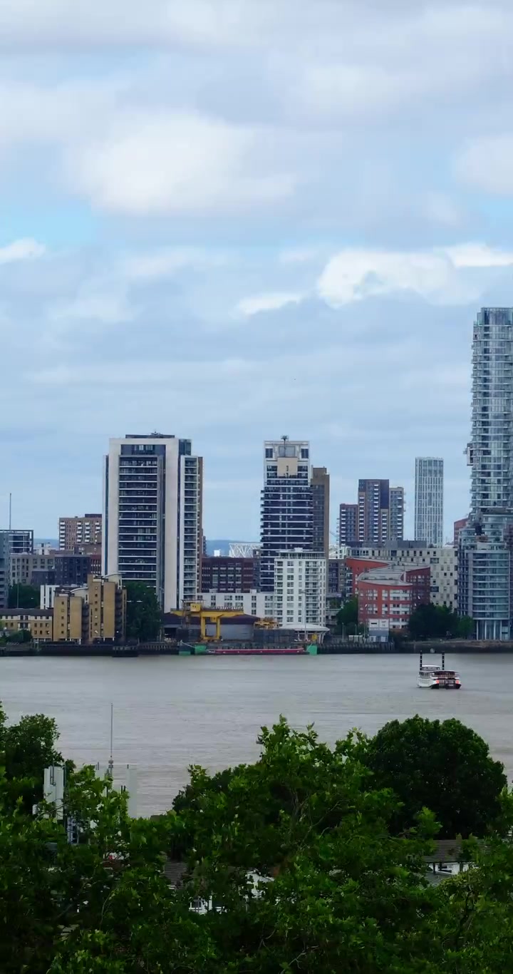 Timelapse vertical do Rio Tâmisa e da área de Canary Wharf, mostrando arranha-céus modernos, arquitetura à beira-mar e nuvens em movimento.