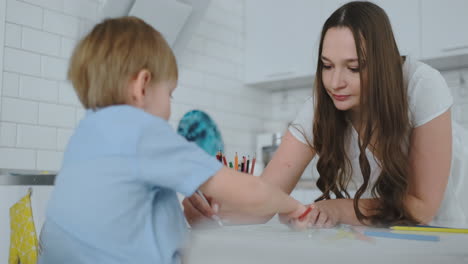 Mamá-Ayuda-A-Dos-Hijos-A-Realizar-La-Tarea-Preescolar-Para-Dibujar-Un-Dibujo-A-Lápiz-Sentado-En-La-Mesa-De-La-Casa