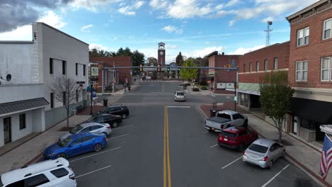 flight on main street on historic american town with clock tower in background. parking cars along uphill road. american flag on storefront in autumn season. drone forward shot.