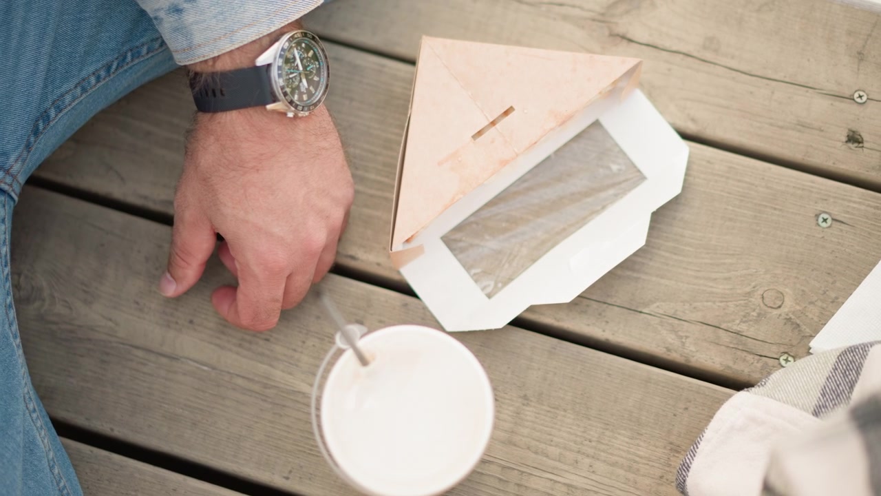 Primo piano della mano di un uomo che poggia su assi di legno accanto a una scatola di cartone triangolare aperta e una tazza bianca con cannuccia. La scena presenta dettagli di un ambiente esterno casual, come la texture del legno e l'illuminazione soffusa