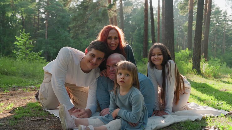 Husband wife and kids smiling while posing closely for family group photo in green forest during sunny day, surrounded by trees and soft light, showing warmth, and joyful outdoor family connection
