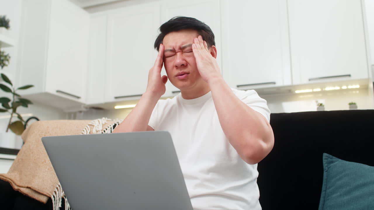 Asian man with laptop feels headache rubs temples closes eyes breathes slowly takes short work break