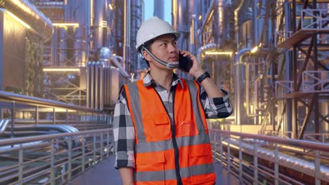asian male engineer with safety helmet talking on smartphone while standing at a vast oil refinery