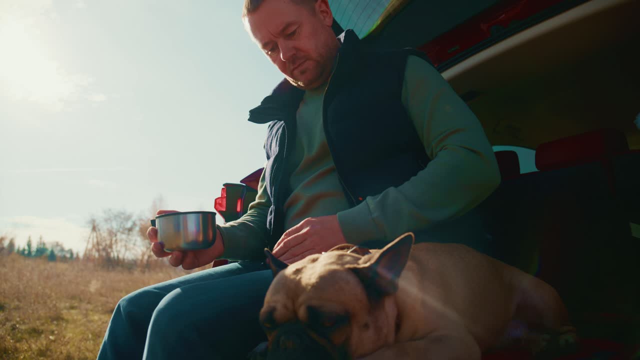 A tranquil moment where a man relaxes in his car's trunk while enjoying the warm sun and comforting presence of his pet dog, creating a serene atmosphere of companionship and peace in nature