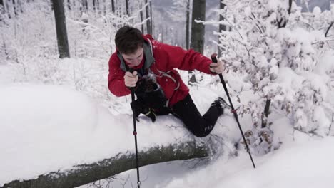 Joven-Excursionista-Masculino-Con-Bastones-De-Senderismo-Trepando-Por-Un-Tronco-De-Madera-Cubierto-De-Nieve-En-El-Bosque