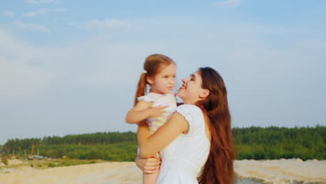 Attractive-Young-Mother-Playing-With-Her-Daughter-Hugging-Sit-In-The-Sand-Against-The-Blue-Sky