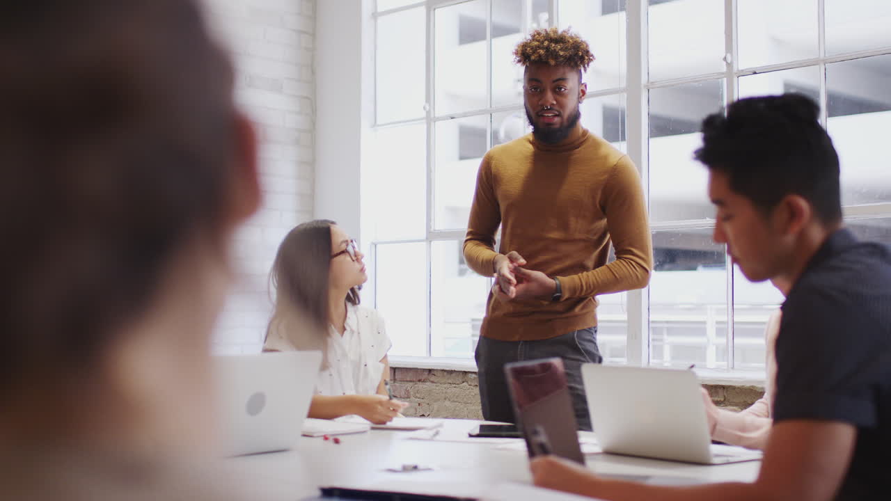 Millennial black man stands addressing his creative colleagues in a meeting room, selective focus
