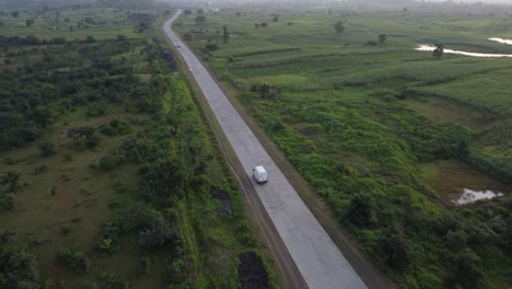 Drone-Siguiendo-Una-Furgoneta-De-Transporte-De-Mercancías-Que-Viaja-Por-Una-Carretera-India-Rural-De-Buena-Calidad-Con-Los-Alrededores-De-Tierras-De-Cultivo-Verdes-Y-Casas-De-Aldea