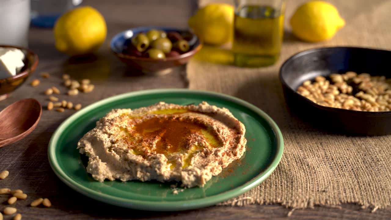 Inquadratura calda e ad angolo basso verso destra che mostra una tavola mediterranea imbandita con olive, pinoli, hummus, feta e ouzo