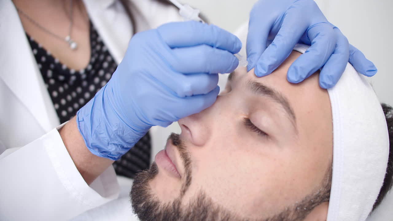 Beautiful slow motion profile shot of a young bearded man receiving a beauty treatment in a dermatology clinic while a female doctor injects him with botox