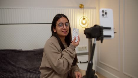 a happy brunette girl in a brown jacket blogs about medicines and pills using a white smartphone in a modern apartment sitting on the bed at home