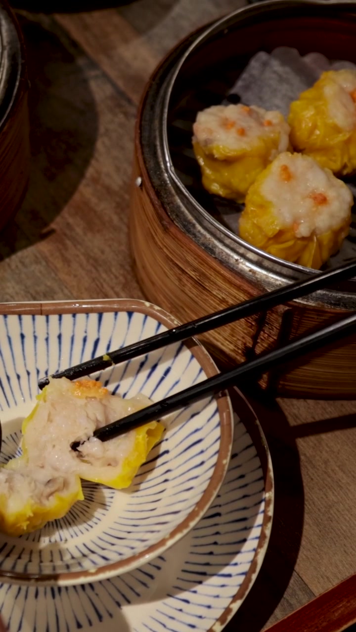 A person uses chopsticks to move a yellow siu mai dumpling into a small bowl.